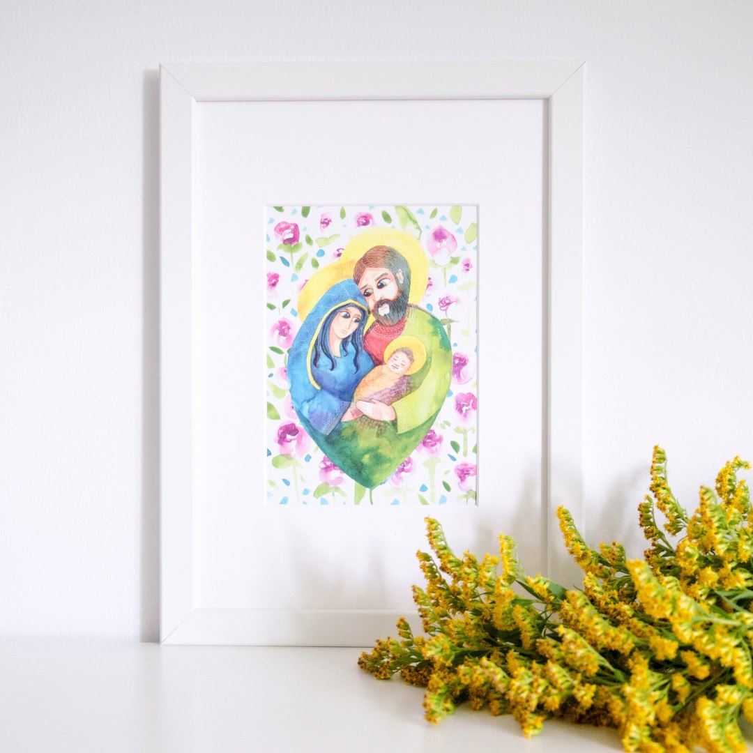 Miniplakat z akwarelowym wizerunkiem Świętej Rodziny. Maryja i Józef przytulają razem małego Jezusa. Otaczają ich kwiaty.  Oprawiony w białe passe-partout i białą prostą ramę.
