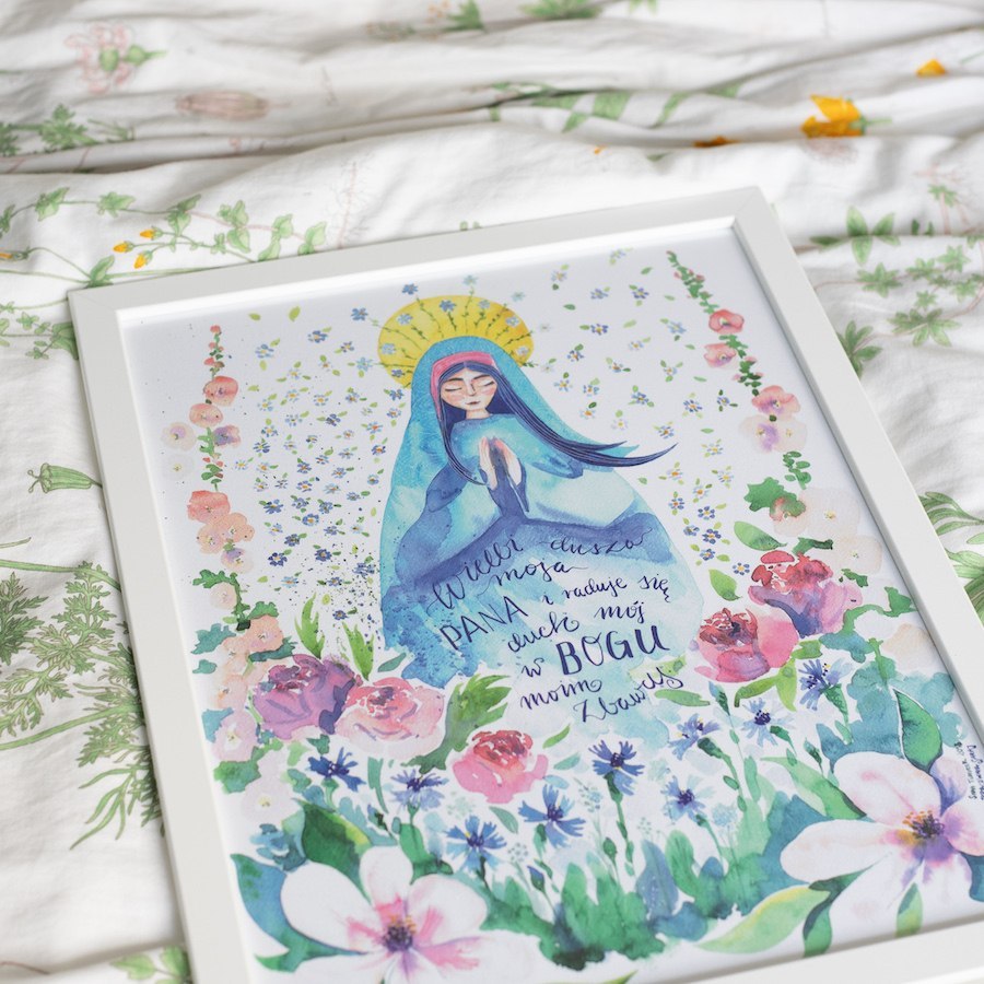 Plakat z modlącą się Maryją. Otaczają ją kwiaty na łące. Oprawiony w białą ramę.