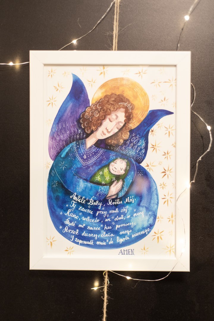 Plakat z ciemnoniebieskim aniołem przytulającym dziecko i modlitwą do Anioła Stróża. Oprawiony w białą prostą ramę.