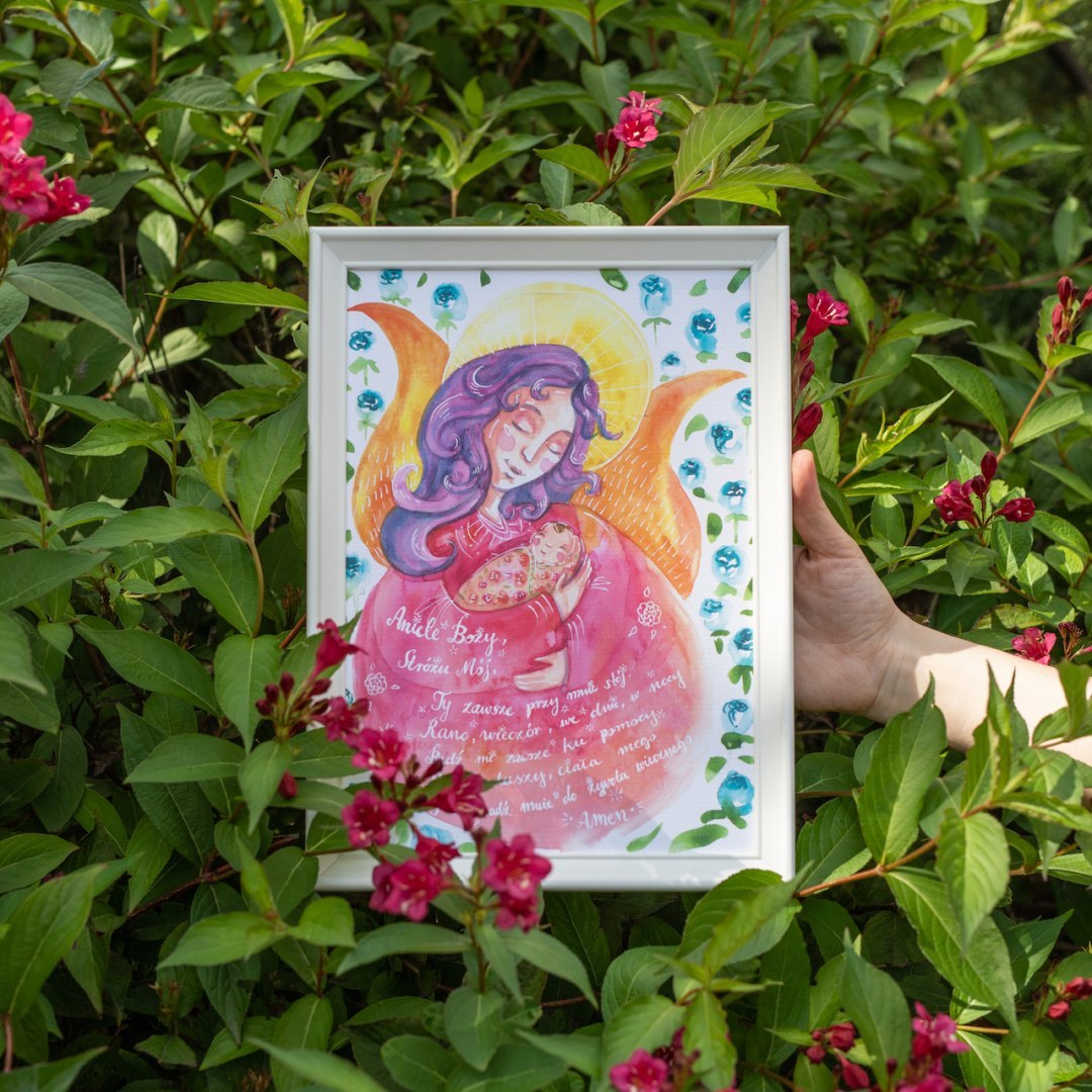 Plakat z różowym aniołem przytulającym dziecko i modlitwą do Anioła Stróża. Oprawiony w białą frezowaną ramę.