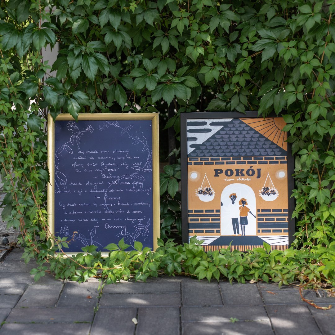 Pomarańczowo-granatowy plakat z parą stojącą przed domem.  Oprawiony w czarną prostą ramę.